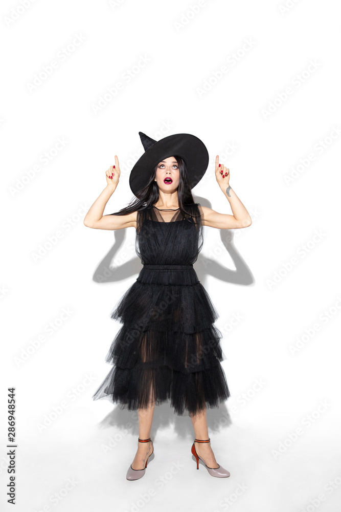 Young brunette woman in black hat and costume on white background. Attractive caucasian female model. Halloween, black friday, cyber monday, sales, autumn concept. Copyspace. Pointing, posing. Young brunette woman in black hat and costume on white background. Attractive caucasian female model. Halloween, black friday, cyber monday, sales, autumn concept. Copyspace. Pointing, posing.