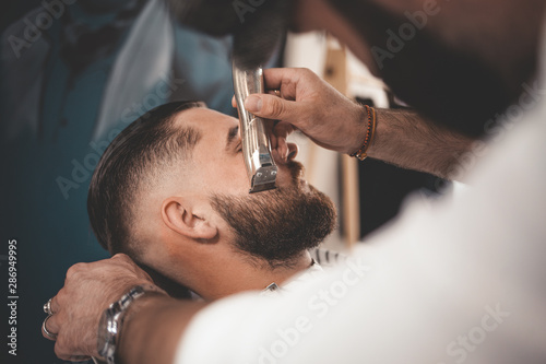Barber makes a beard cut for a client. Hipster client visited barbershop 