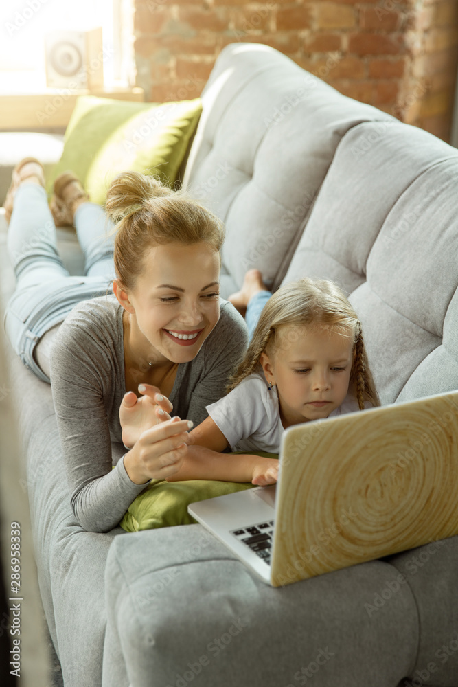 Female caucasian teacher and little girl, or mom and daughter. Homeschooling. Lying on the sofa and using laptop for knowledges getting while lesson is. Education, school, studying concept. Female caucasian teacher and little girl, or mom and daughter. Homeschooling. Lying on the sofa and using laptop for knowledges getting while lesson is. Education, school, studying concept.
