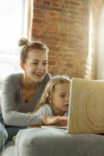 Female caucasian teacher and little girl, or mom and daughter. Homeschooling. Lying on the sofa and using laptop for knowledges getting while lesson is. Education, school, studying concept.