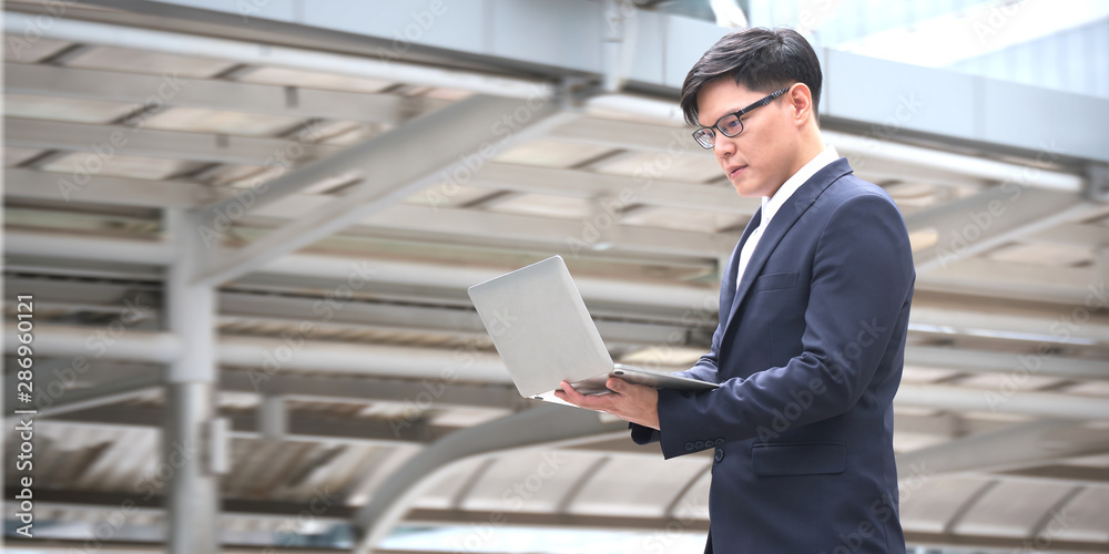 Asian Businessman wearing stylish blue suit and glasses is working on a laptop while standing outdoors at the walkway in Business district with high buildings. Asian Businessman wearing stylish blue suit and glasses is working on a laptop while standing outdoors at the walkway in Business district with high buildings.