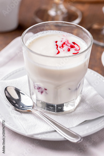 glass of milkshake with berry syrup close-up, white saucer, teaspoon and linen napkin on table 