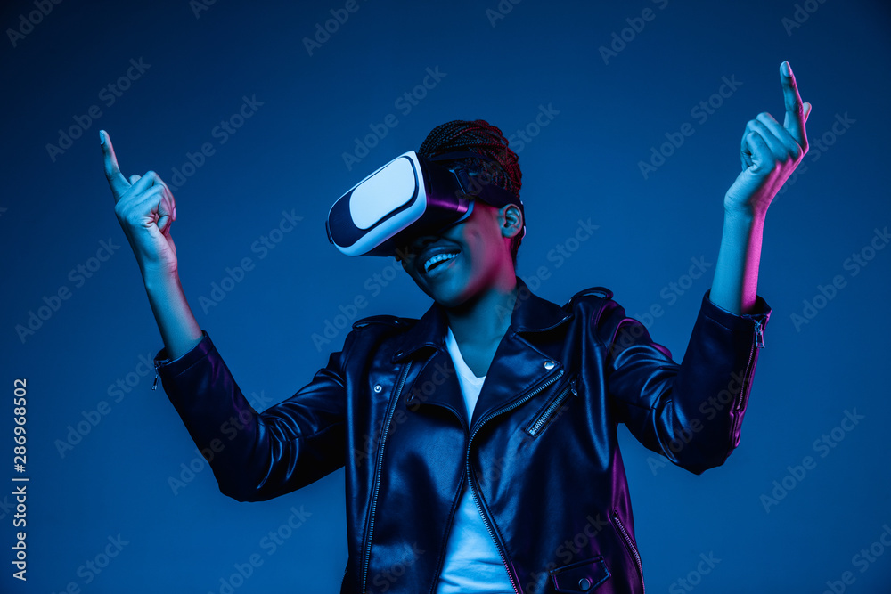 Portrait of young african-american woman's playing in VR-glasses in neon light on blue background. Concept of human emotions, facial expression, modern gadgets and technologies. Look shooting