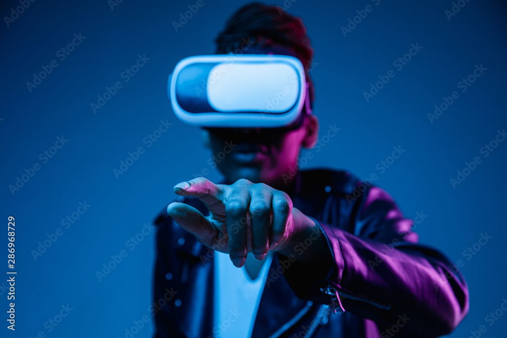 Portrait of young african-american woman's playing in VR-glasses in neon light on blue background. Concept of human emotions, facial expression, modern gadgets and technologies. Touches empty bar. Portrait of young african-american woman's playing in VR-glasses in neon light on blue background. Concept of human emotions, facial expression, modern gadgets and technologies. Touches empty bar.