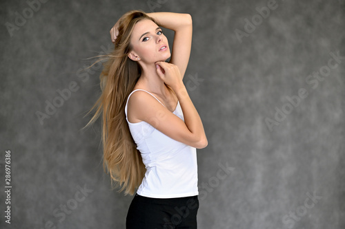 Portrait of a beautiful pretty girl student in a white T-shirt with long curly hair on a gray background. Beauty and brightness. Shows hands to the side, smiles with different emotions.