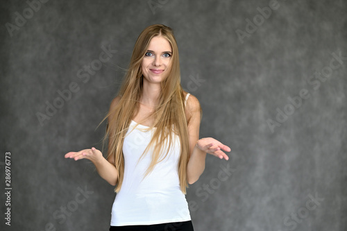 Portrait of a beautiful pretty girl student in a white T-shirt with long curly hair on a gray background. Beauty and brightness. Shows hands to the side, smiles with different emotions.