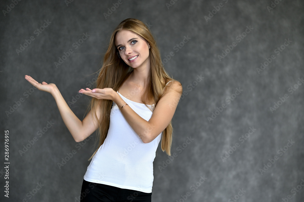 Portrait of a beautiful pretty girl student in a white T-shirt with long curly hair on a gray background. Beauty and brightness. Shows hands to the side, smiles with different emotions. Portrait of a beautiful pretty girl student in a white T-shirt with long curly hair on a gray background. Beauty and brightness. Shows hands to the side, smiles with different emotions.