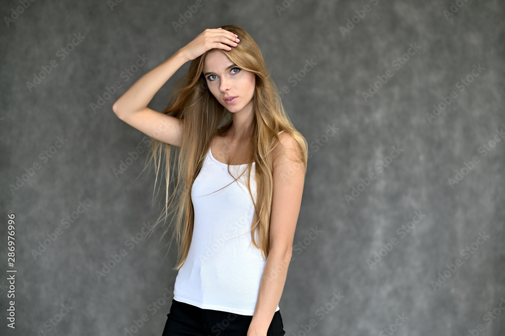 Portrait of a beautiful pretty girl student in a white T-shirt with long curly hair on a gray background. Beauty and brightness. Shows hands to the side, smiles with different emotions. Portrait of a beautiful pretty girl student in a white T-shirt with long curly hair on a gray background. Beauty and brightness. Shows hands to the side, smiles with different emotions.
