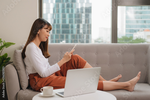 Young woman in casual clothes sitting on sofa at cozy home interior. Technology and communication concept.