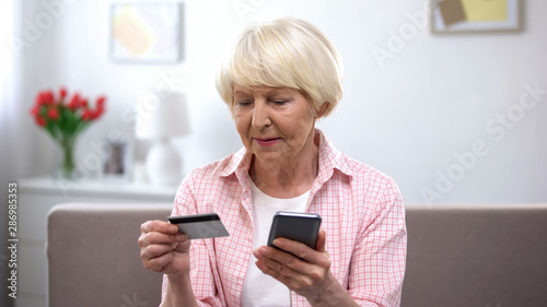 Confused aged woman with credit card and smartphone trying to pay bills online