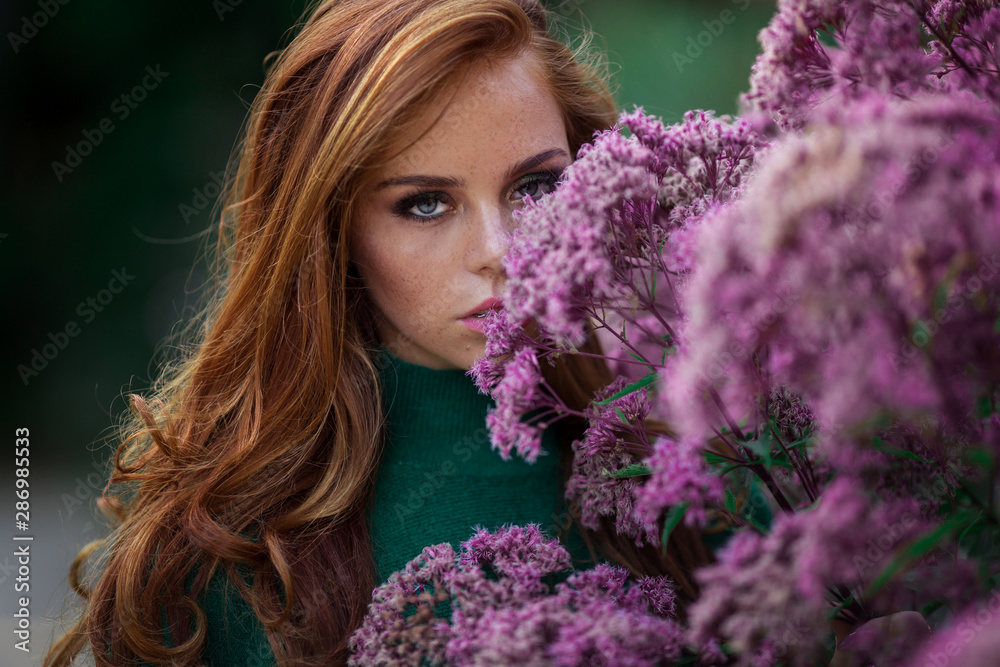 Sunshine young smiling woman with red curly hair is wearing green sweater and hat in autumn park near purple bush of flowers. Sunshine young smiling woman with red curly hair is wearing green sweater and hat in autumn park near purple bush of flowers.