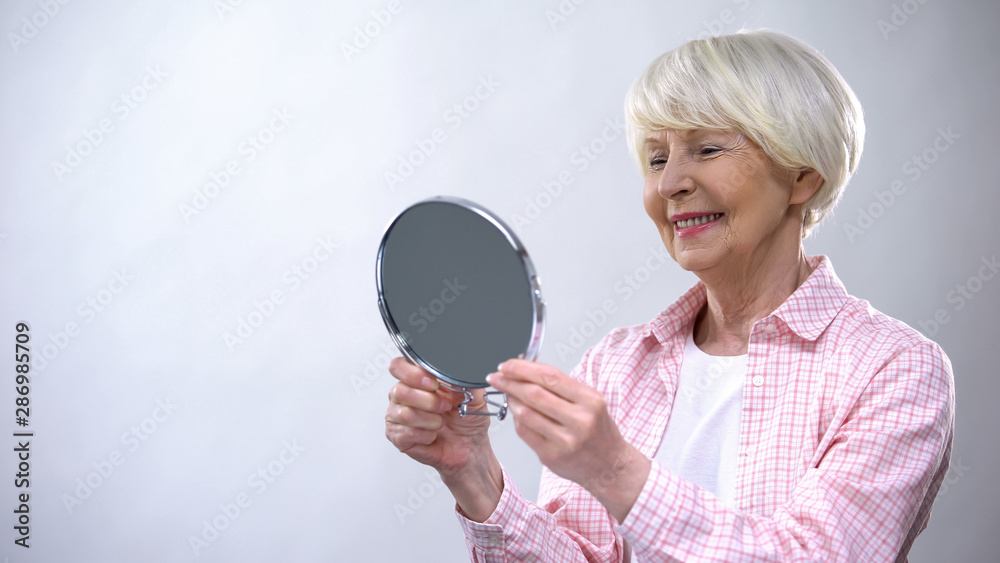 Smiling aged woman looking at mirror reflection, enjoying beauty, appearance Smiling aged woman looking at mirror reflection, enjoying beauty, appearance
