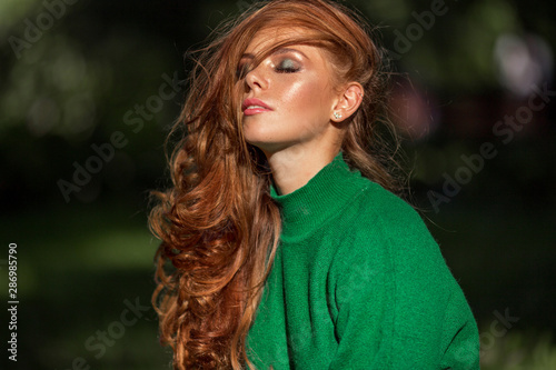 Sunshine young smiling woman with red curly hair is wearing green sweater in autumn park.