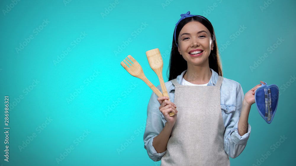 Pretty housewife in apron holding kitchenware and smiling at camera, cooking Pretty housewife in apron holding kitchenware and smiling at camera, cooking