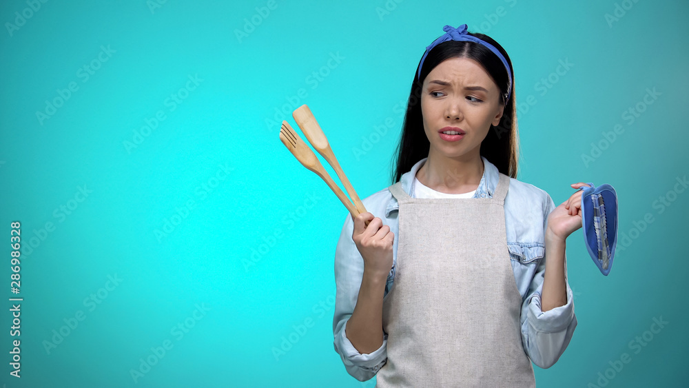 Incompetent housewife in apron holding kitchenware and shrugging shoulders Incompetent housewife in apron holding kitchenware and shrugging shoulders