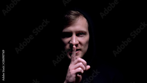 Billede på lærred Hooded man with spooky grin holding finger near lips showing shh sign, threat