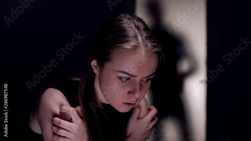 Scared crying woman sitting alone in room, cruel tyrant husband shadow outside