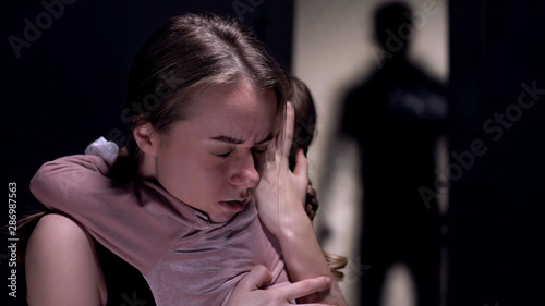 Fotografie Frightened mother hugging daughter, felon shadow entering room, crime witness