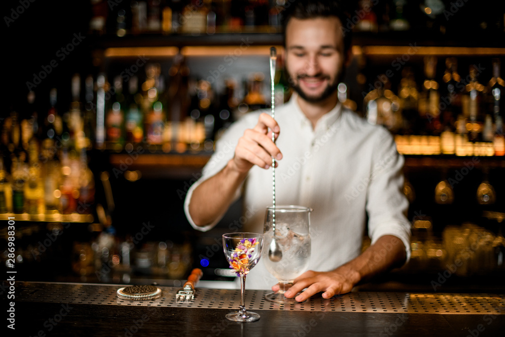 Smiling bartender stirring a cocktail with a spoon in the focus foreground of flower decorated glass