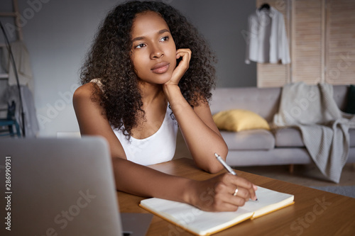 Occupation, work, technology and connection. Thoughtful pensive young dark skinned woman with voluminous hair using laptop for remote work, writing down in copybook, trying to recollect something