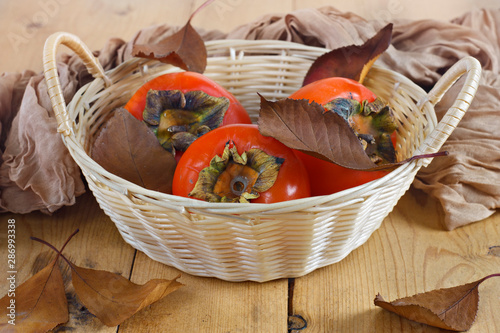 Autumn fruits. Persimmons in a wicker basket on a wooden board on a wooden background. Fallen leaves.