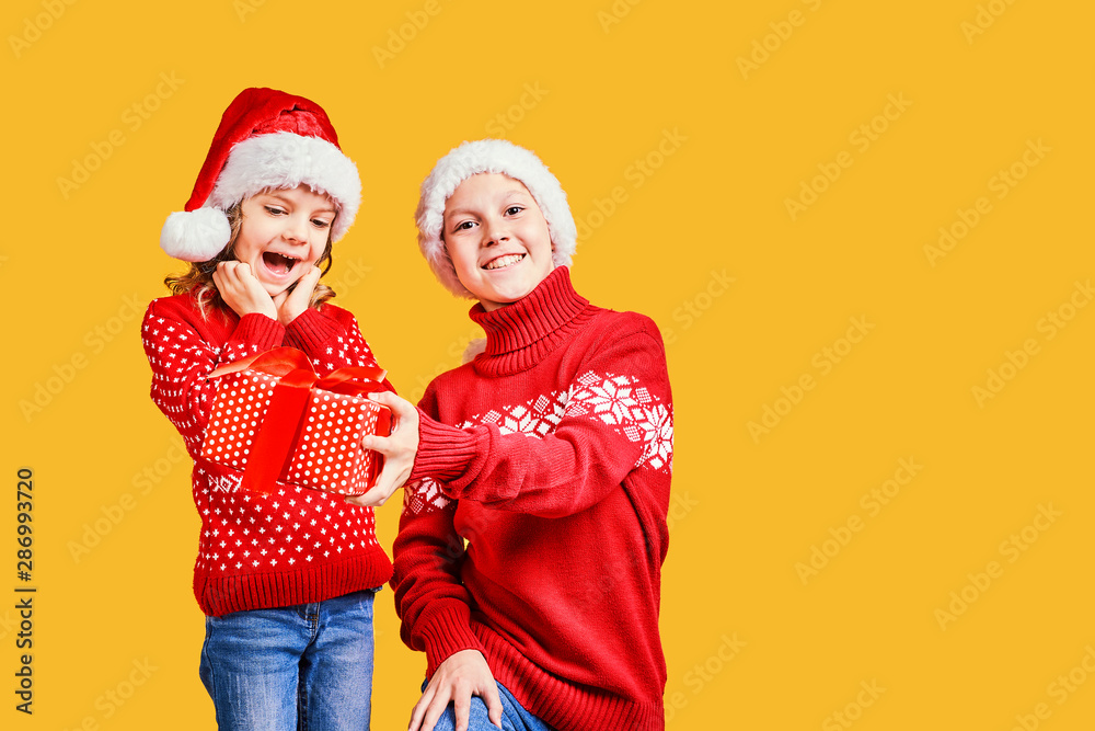 Fototapeta premium Happy boy in Santa hat giving Christmas gift box to excited girl in red sweater with deer on yellow background