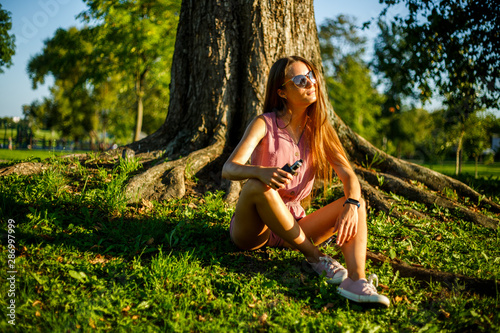 Vape teenager. Young pretty sporty caucasian girl in a pink jumpsuit and sunglasses smoking an electronic cigarette in the park on a sunny day in summer. Bad habit. Vaping activity.