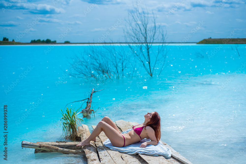 A beautiful red-haired woman in a seductive crimson bikini sits in front of an unusual turquoise lake. A miracle of nature, bright blue clear water. Ash dump, poisoned pond.