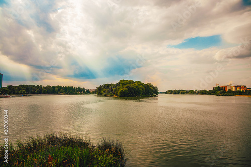 Eastern European city outskirts scenic view of island in lake surrounded by green park space with buildings on background and sun rays light through white and gray clouds 