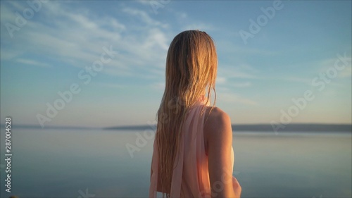  beautiful women in a light pink dress walking along the beach at dawn. Good morning and relaxation. Young beautiful girl stands in the water in a light pink dress on the background of the dawn.