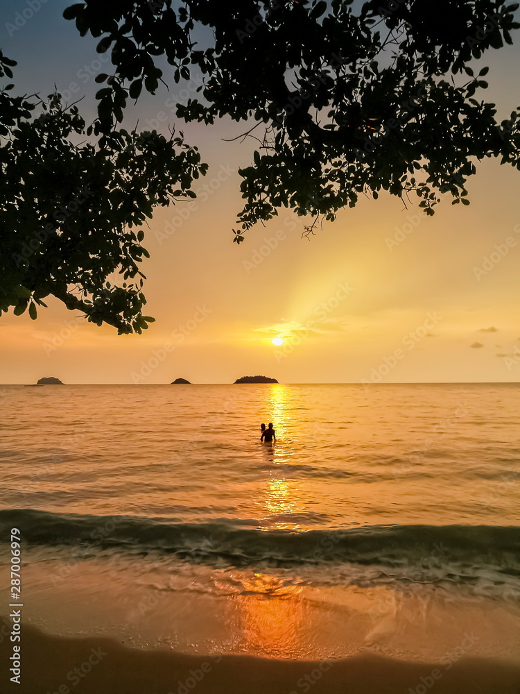 view seaside evening under tree branch of couple tourist playing in the sea with golden sun light background, sunset at Kai Bae Beach, Koh Chang, Trat, east of Thailand.
