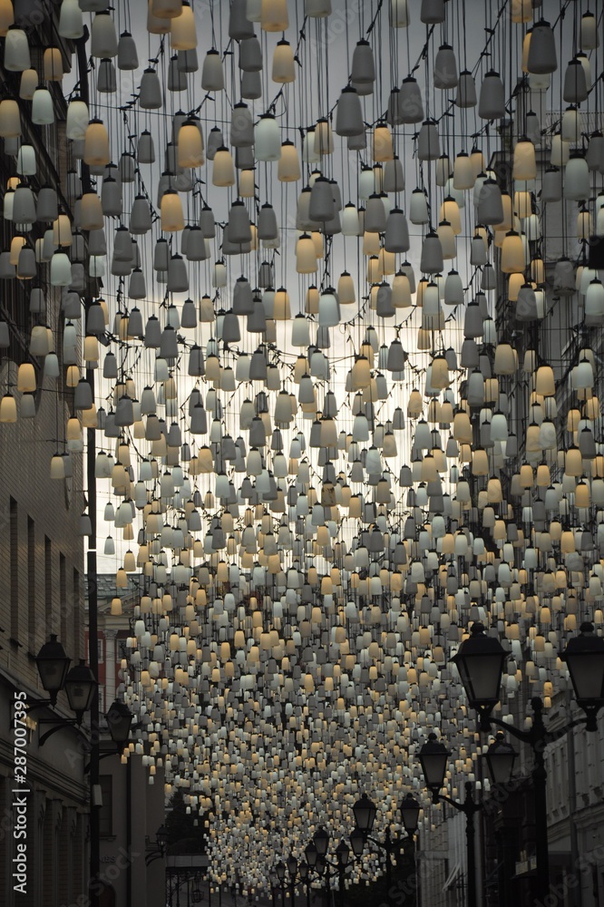 Obraz premium An alley of Moscow with lanterns standing and fortified on the walls of houses and yellow and white bulbs hanging in the sky.