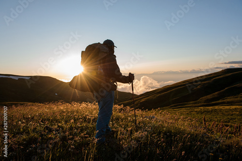 Rear view man standing on the hill field with hiking backpack and sticks looking at the sun