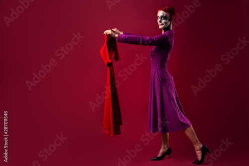 A girl with clown makeup in a purple outfit holds a red dress in her hands. The concept of shopping and choosing clothes