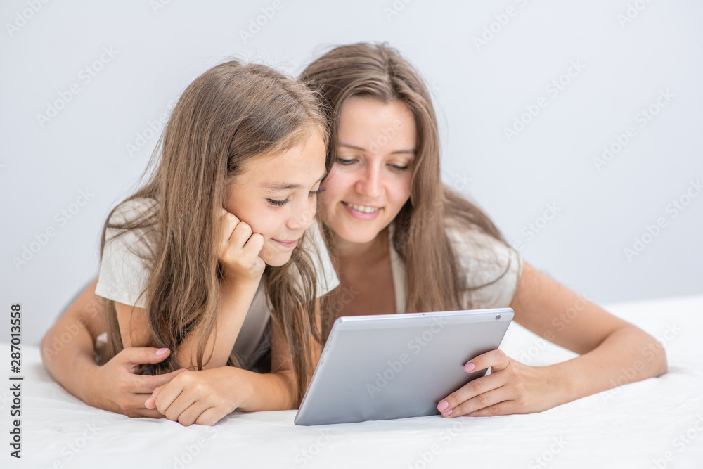 Happy family at home. Beautiful young woman hugging her little daughter and using a digital tablet and smiling while lying in bed at home
