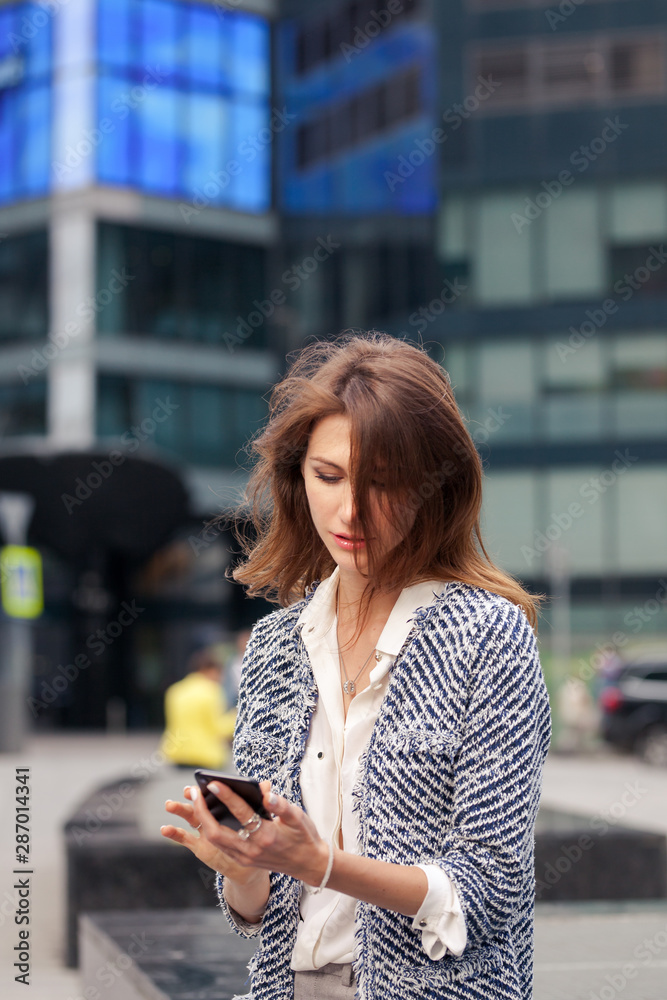Fototapeta premium beautiful young business brunette woman dressed in elegant clothes sits on a bench on a street in the city center and talking on a cell phone. Glass building in the background