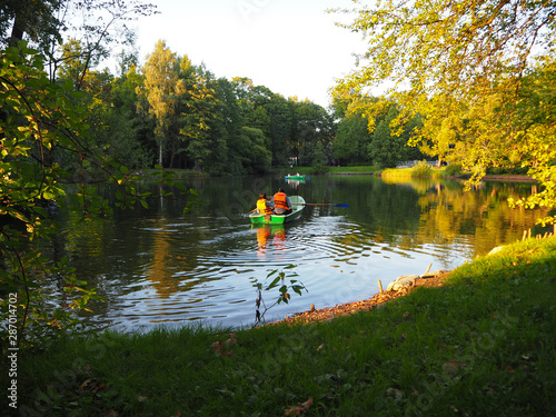 Autumn park landscape with people boating in a lake and the green foliage of the trees start to turn yellow.