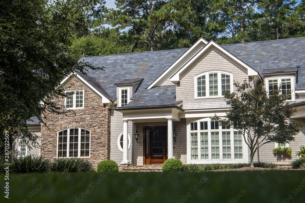 Front view of large estate home in the south with a gravel driveway and lots of windows. house made of brick, stone and clapboard in a cape cod style. and a triple garage with curb appeal