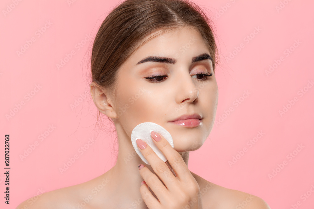 Beautiful model applyes cosmetic product to skin with white cotton sponge. Model looks at the camera isolated over pink background. Beautiful model applyes cosmetic product to skin with white cotton sponge. Model looks at the camera isolated over pink background.