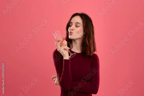 Excited pretty brunette girl holding wineglass of sparkling champagne standing isolated on a dark pink background and smiling at the camera.