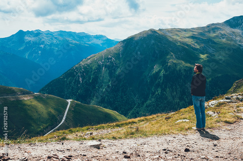 Tourist man standing on the top of Alp mountains, looking at the valley beyond the mountain pass, enjoying the beautiful landscape. Travel, adventure, freedom, challenge, hiking, vacation concept