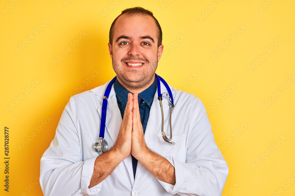 Young doctor man wearing coat and stethoscope standing over isolated yellow background praying with hands together asking for forgiveness smiling confident. Young doctor man wearing coat and stethoscope standing over isolated yellow background praying with hands together asking for forgiveness smiling confident.