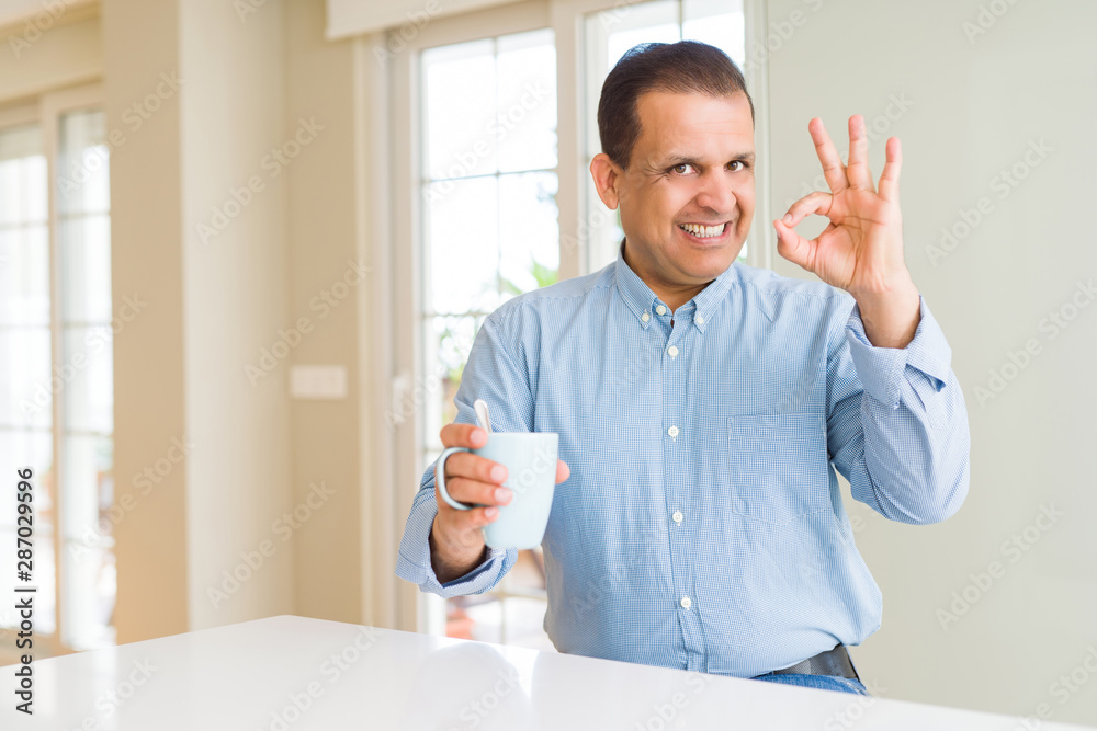Middle age man drinking coffee in the morning at home doing ok sign with fingers, excellent symbol