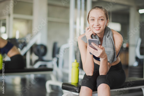 Young woman take a rest after workout at gym which looking at camera and playing a smart phone. Woman sitting and searching in smartphone at the fitness room near big mirror which smiling and happy.
