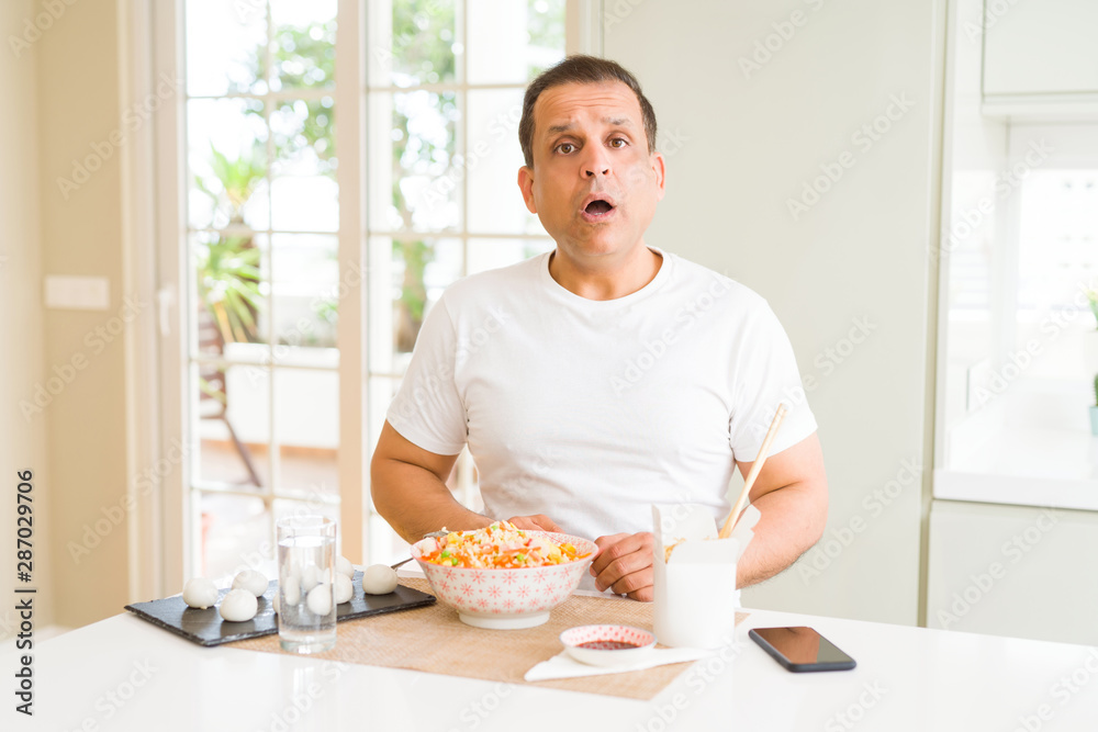 Middle age man eating asian food with chopsticks at home afraid and shocked with surprise expression, fear and excited face. Middle age man eating asian food with chopsticks at home afraid and shocked with surprise expression, fear and excited face.