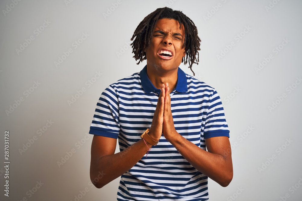 Afro man with dreadlocks wearing striped blue polo standing over isolated white background begging and praying with hands together with hope expression on face very emotional and worried. Afro man with dreadlocks wearing striped blue polo standing over isolated white background begging and praying with hands together with hope expression on face very emotional and worried.