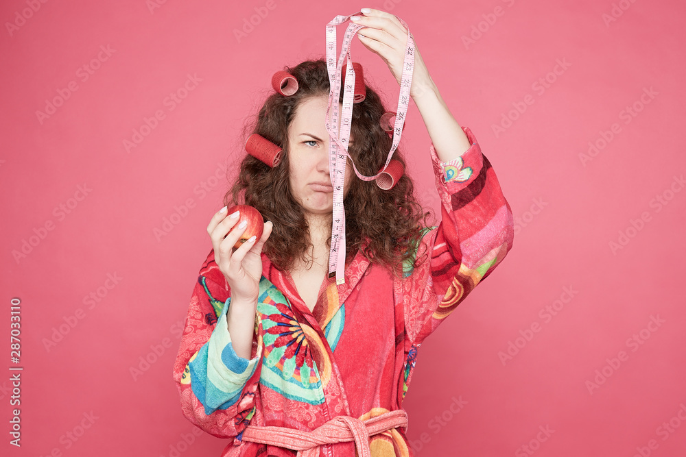 Depressive beautiful emotional lady looks with fearful expression at  measuring tape, holds fresh red apple in hand, dressed in casual bathrobe,  models against pink background with blank copy space. Stock Photo