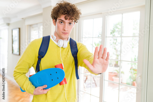 Young student man wearing headphones and backpack holding skateboard with open hand doing stop sign with serious and confident expression, defense gesture © Krakenimages.com