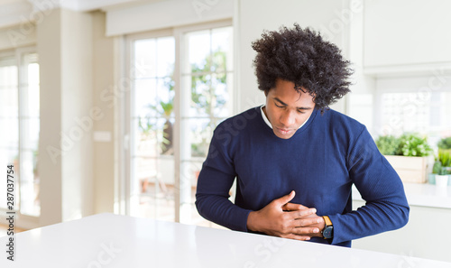 Young african american man wearing casual sweater sitting at home with hand on stomach because indigestion, painful illness feeling unwell. Ache concept.