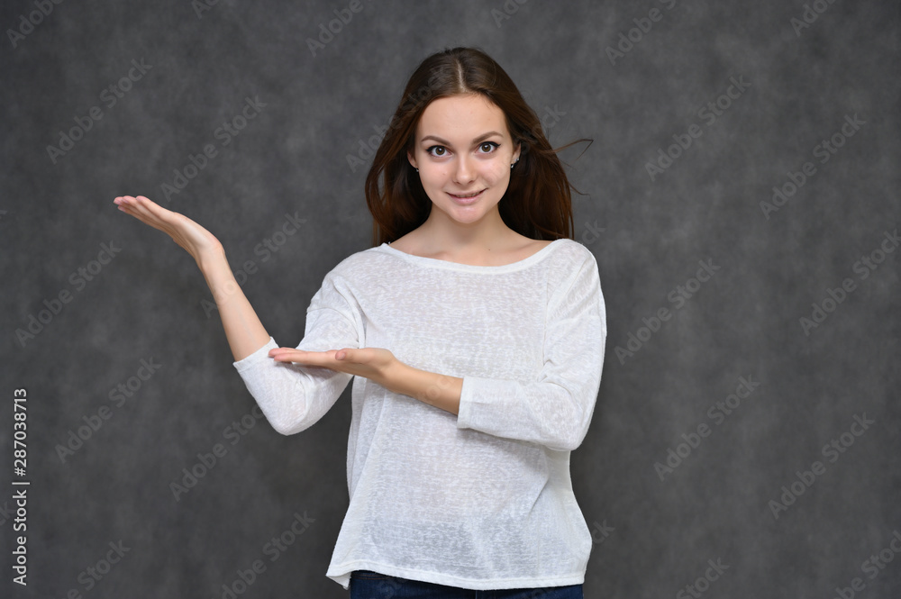 Portrait of a cute brunette girl, a young woman with beautiful curly hair in a white sweater on a gray background. Smiling, talking with emotions, showing hands to the sides. Portrait of a cute brunette girl, a young woman with beautiful curly hair in a white sweater on a gray background. Smiling, talking with emotions, showing hands to the sides.